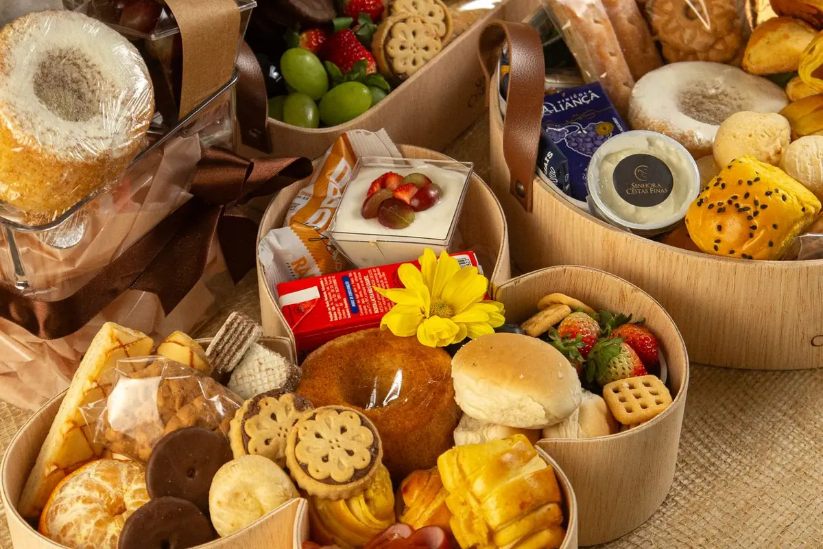 Mesa de café da manhã com três cestas de madeira cheias de pães, frutas, bolos e biscoitos sobre juta