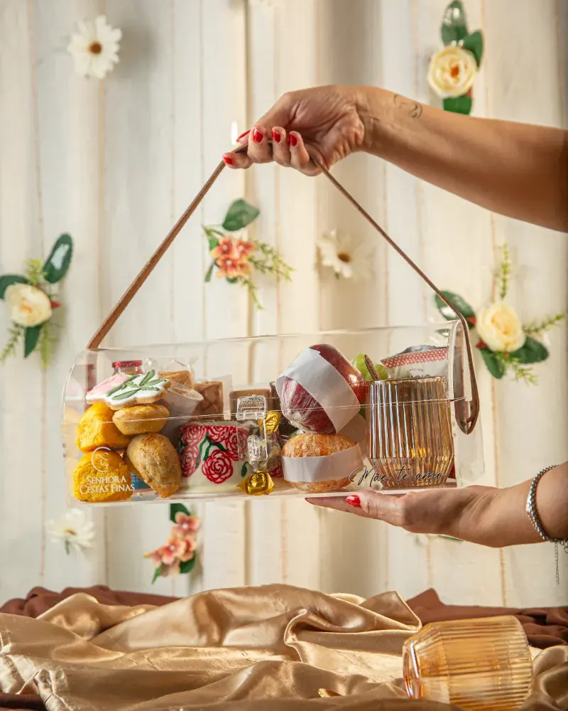 Baú de acrílico da Luxury Breakfast segurado pela alça de couro, com pães, frutas e xícara, diante de uma parede de flores