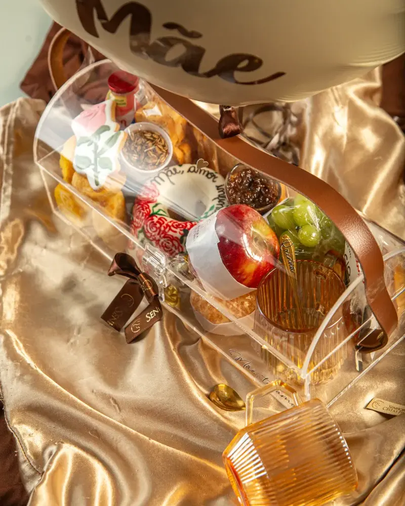 Itens de recordação da Luxury Breakfast, como a xícara de vidro e a colher, ao lado do baú de acrílico sobre tecido dourado