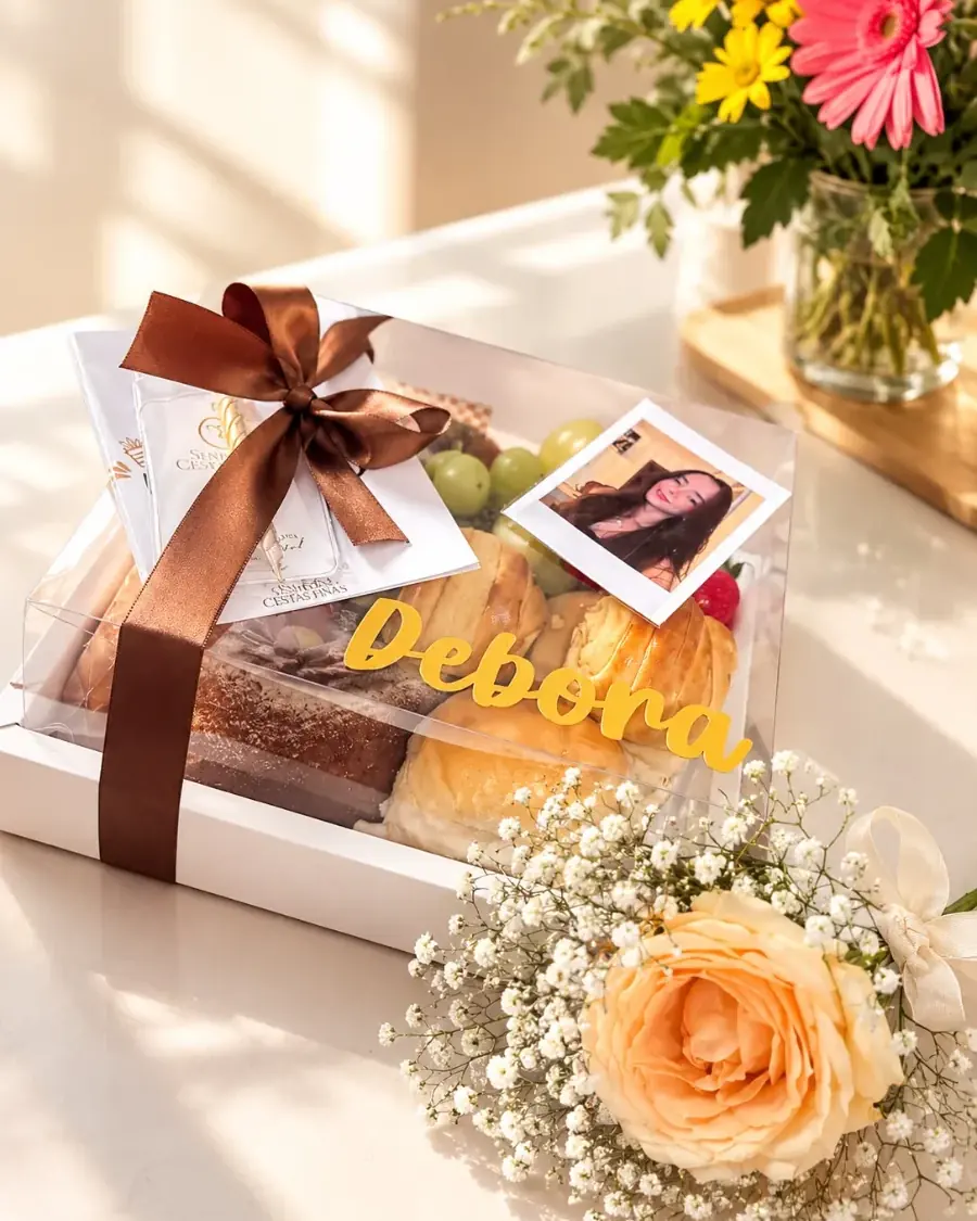 Cesta Mimo Day em box de acetato transparente com laço marrom, pães, bolo, polaroide e um arranjo de flores ao lado