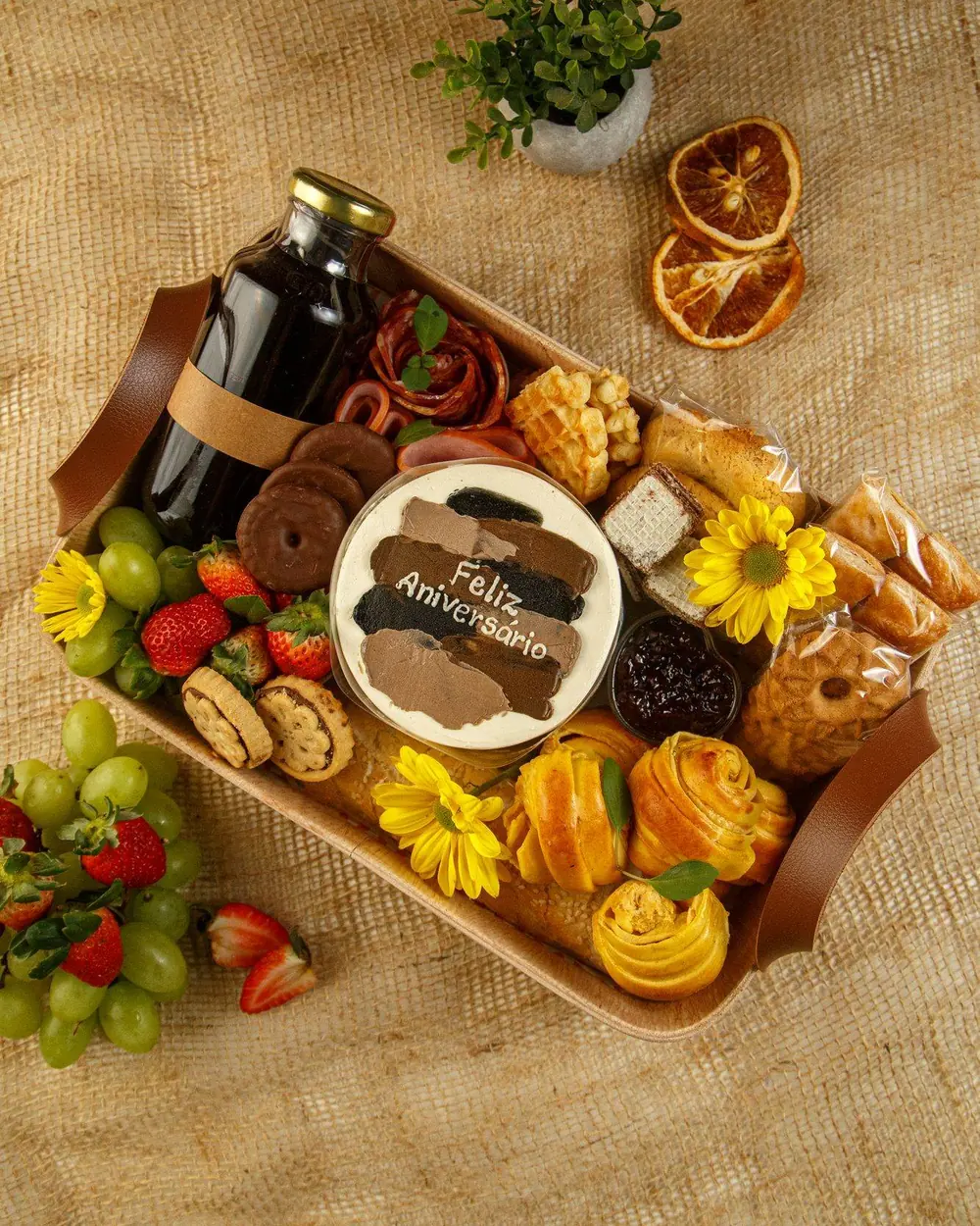 Cesta Morning Elegance vista de cima, em bandeja com alças de couro, com suco, bolo Feliz Aniversário, frios, frutas, pães e biscoitos sobre juta