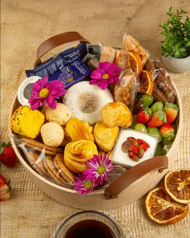 Cesta redonda de café da manhã Morning Soft, em madeira clara com alças de couro, com pão de queijo, croissant, bolo polvilhado com açúcar, biscoitos, morangos, uvas verdes, um potinho de iogurte com frutas, flores decorativas e rodelas de laranja desidratada, sobre um tecido de juta.