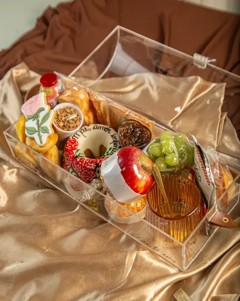 Baú de acrílico da Luxury Breakfast visto de cima, com frutas, pães, doces e xícara sobre tecido dourado