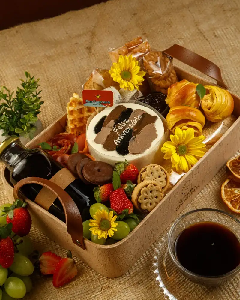 Cesta Morning Elegance com alças de couro, bolo de aniversário, croissants, frutas e uma xícara de café ao lado