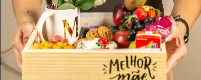 Cesta de madeira com a frase Melhor mãe, caneca com a inicial M, frutas e biscoitos, segurada por uma profissional da Senhora Cestas Finas
