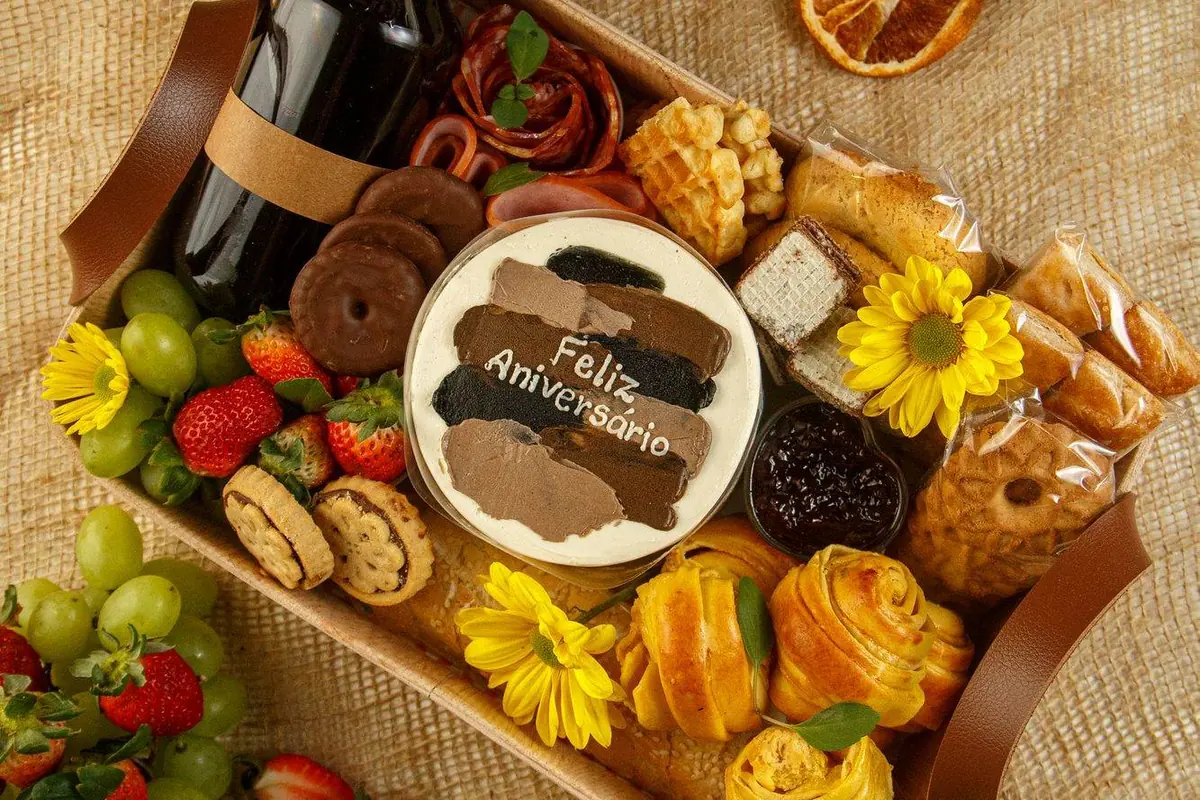 Cesta de aniversário vista de cima, com bolo Feliz Aniversário, suco, frios, frutas e pães em bandeja com alças de couro sobre juta