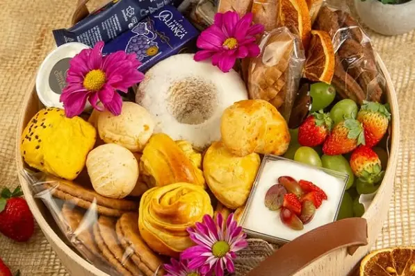 Cesta redonda de madeira com alças de couro, rosca, pães, morangos, uvas, flores e uma xícara de café