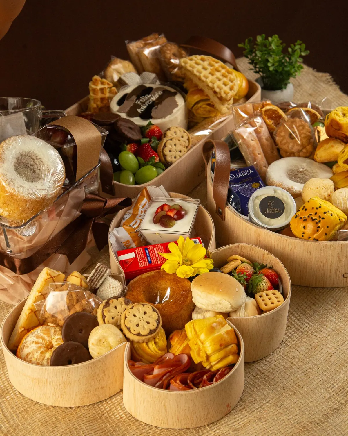 Três cestas de madeira com pães, frutas, bolos, biscoitos e frios sobre juta, com uma caneca de vidro ao fundo
