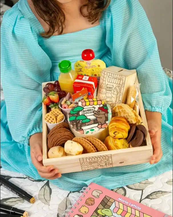 Menina de vestido azul segurando a cesta Happy Box Kids, em caixa de madeira com bento cake temático, suco, biscoitos, pães e cofrinho