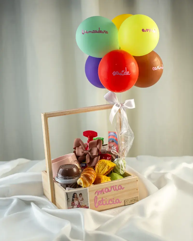 Cesta infantil de madeira com balões coloridos amarrados, sobre tecido branco