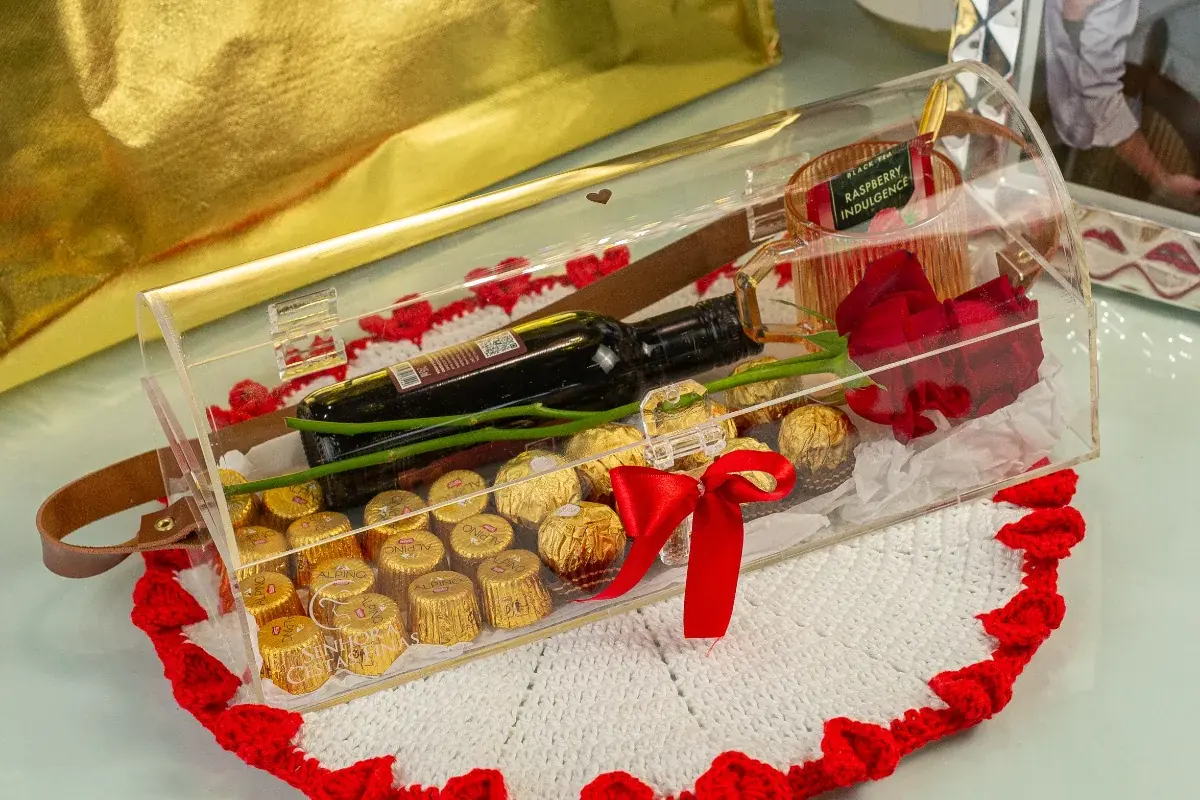 Baú de acrílico da Forever Us Love com vinho, chocolates, rosa vermelha e laço vermelho sobre um sousplat de crochê