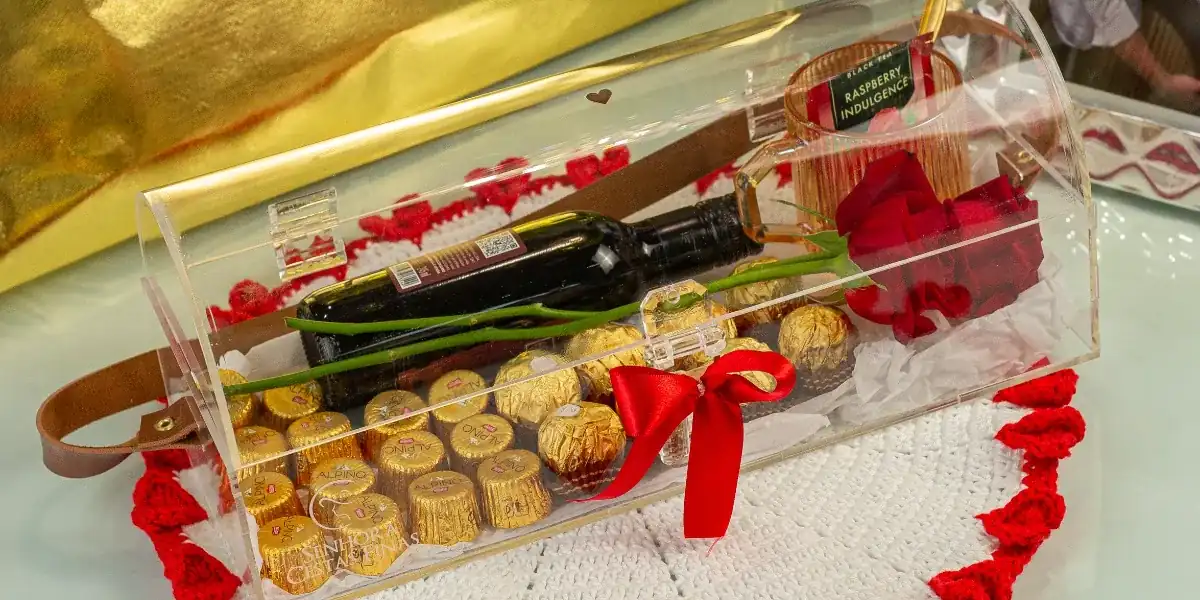 Baú de acrílico da Forever Us Love com vinho, chocolates, rosa vermelha e laço vermelho sobre um sousplat de crochê